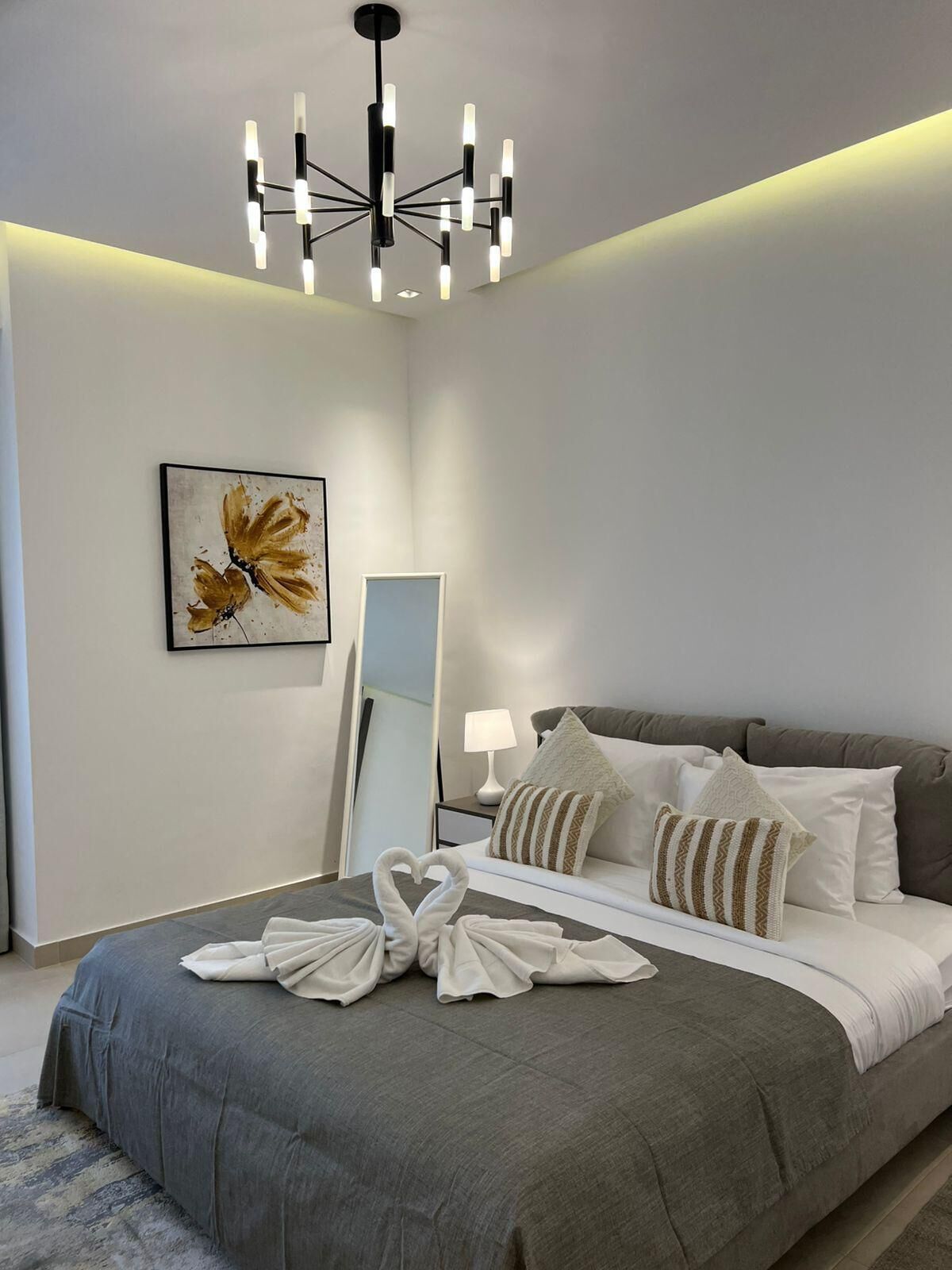 Un dormitorio moderno con una cama gris, almohadas blancas, un espejo, un cuadro y una araña.