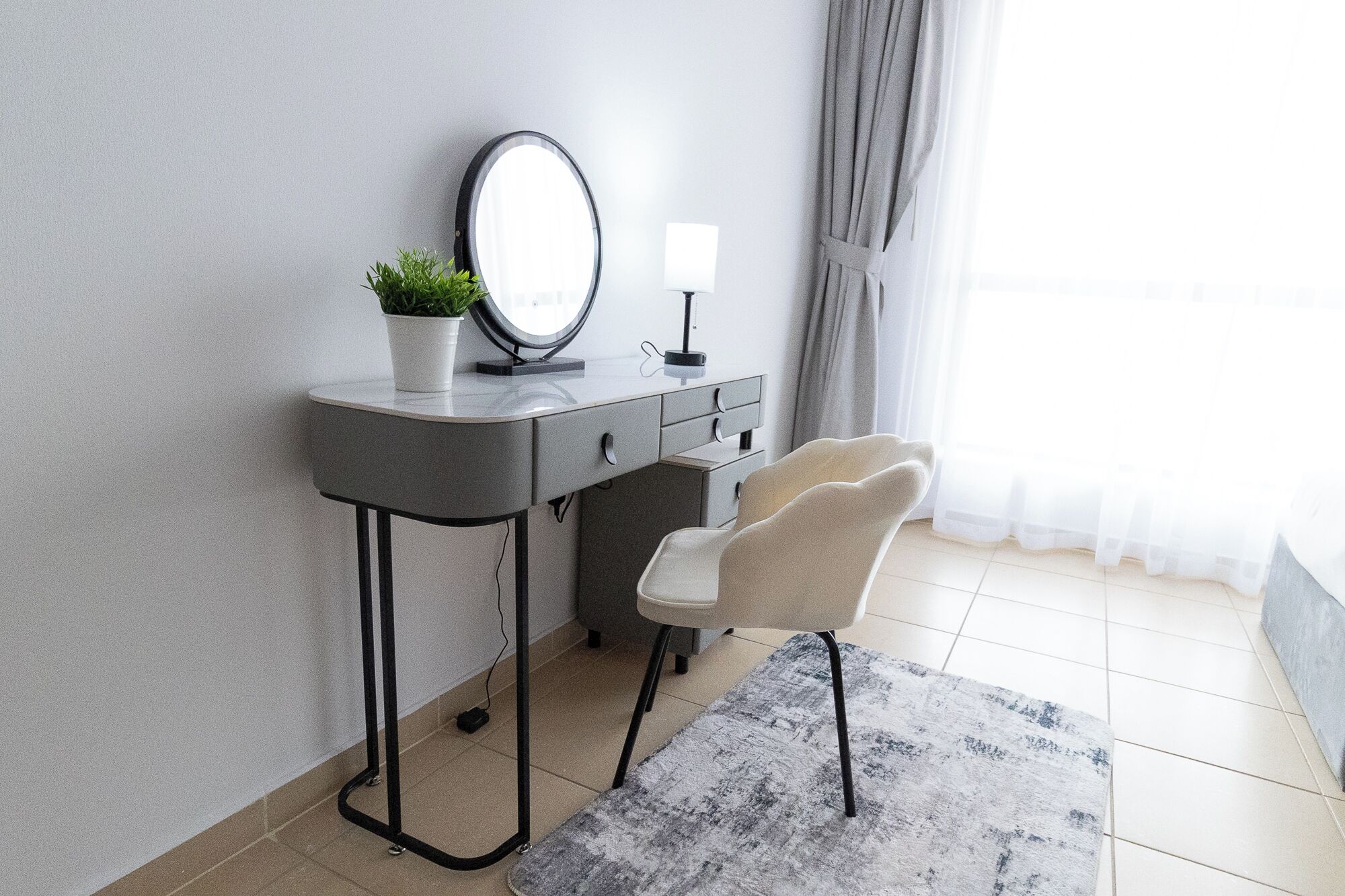 A modern bedroom with a grey vanity, a round mirror, a small potted plant, a white bedside lamp, a grey chair, and a grey rug.