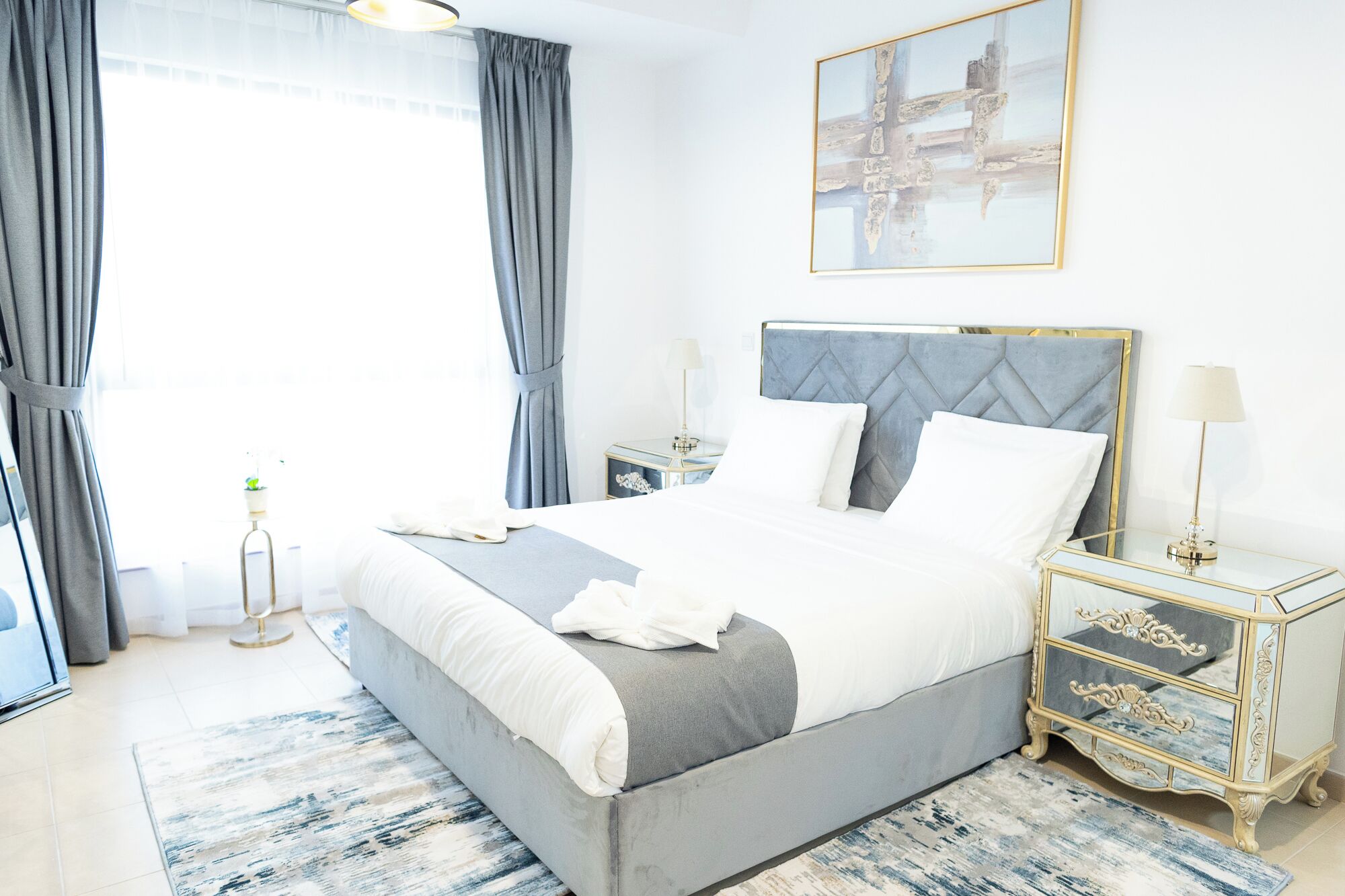 A modern hotel room with a large bed, two bedside tables, a nightstand, a window with curtains, and a painting on the wall.