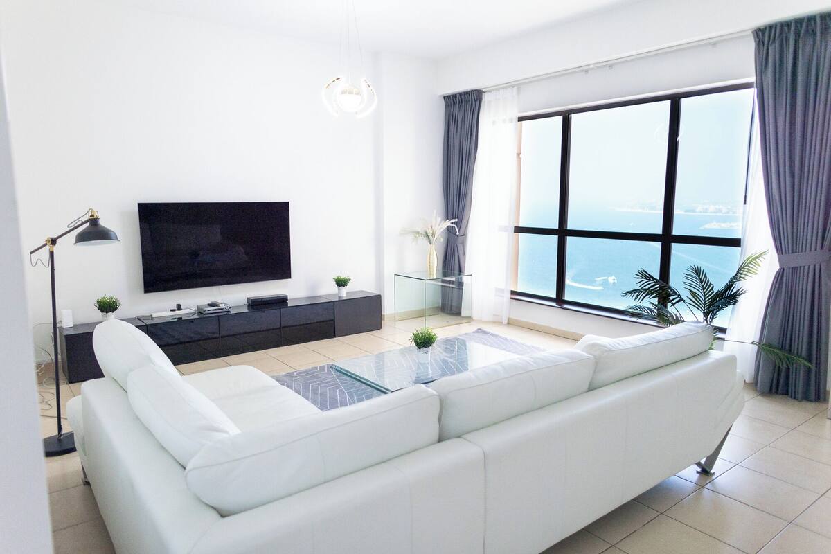 A modern living room with a white sofa, a flat-screen TV, a glass coffee table, and a view of the sea through large windows.