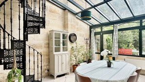 Une pièce spacieuse avec un plafond haut, un patio vitré, un escalier décoratif, une table à manger avec des chaises et un buffet.