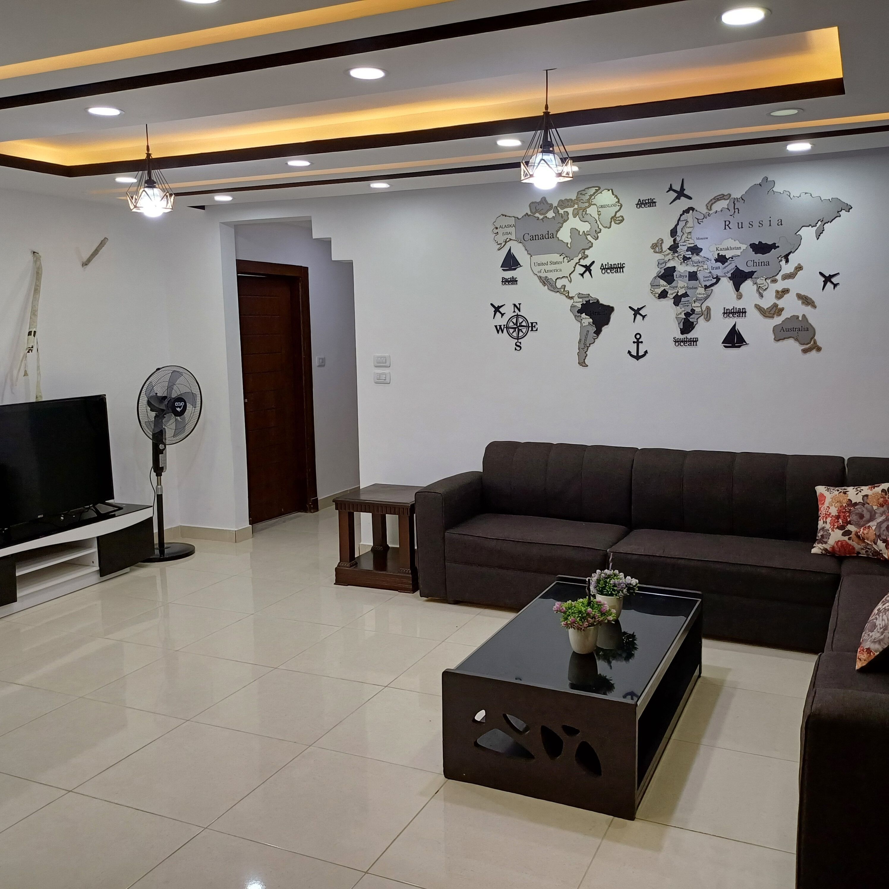 A modern living room with a flat-screen TV, a dark brown sofa, a coffee table with a floral centerpiece, and a world map on the wall.