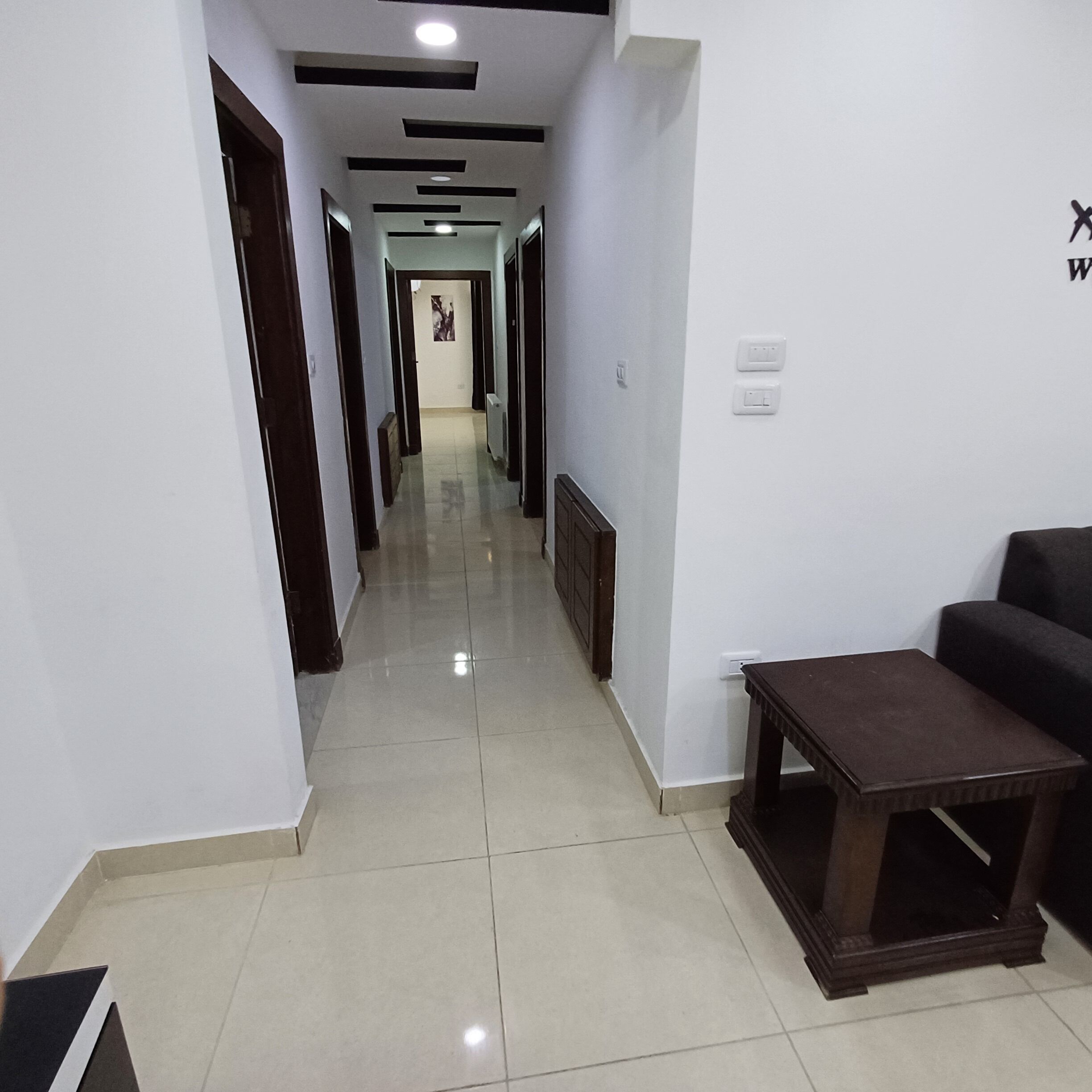 A hallway with a tiled floor, a dark wooden table, and a white wall with a power outlet.