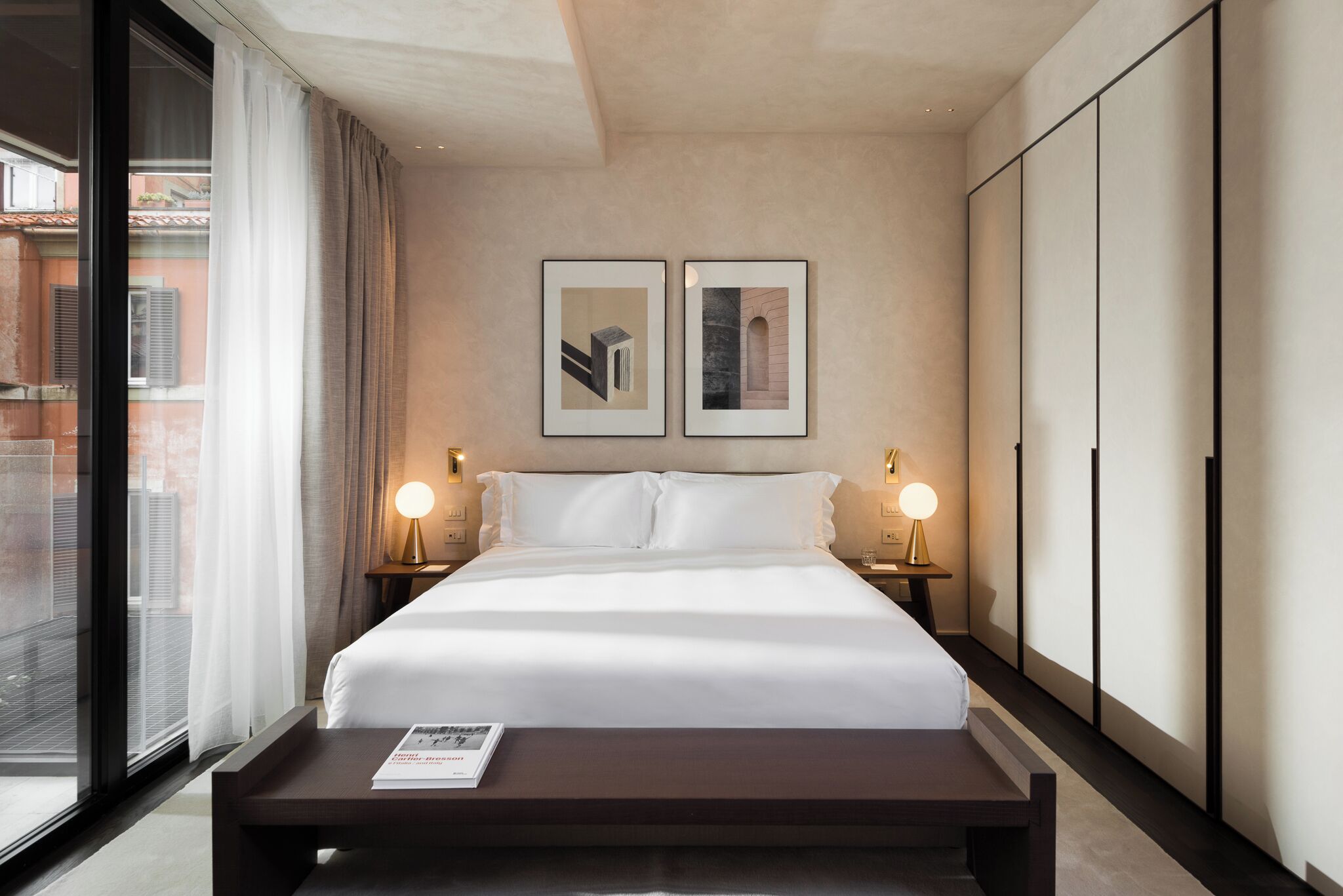 A hotel room with a large bed, two bedside tables with lamps, a window with curtains, and two framed pictures on the wall.