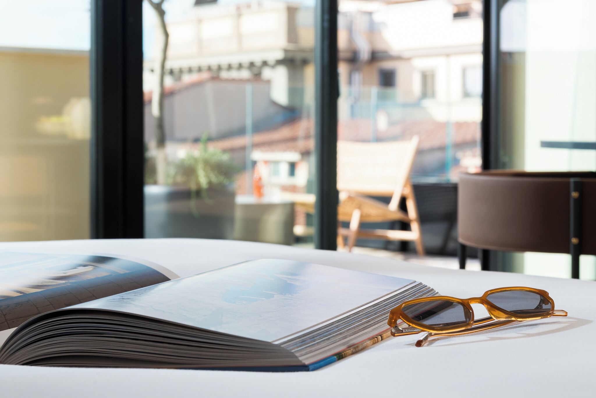 A book and glasses on a table with a view of buildings through a window.