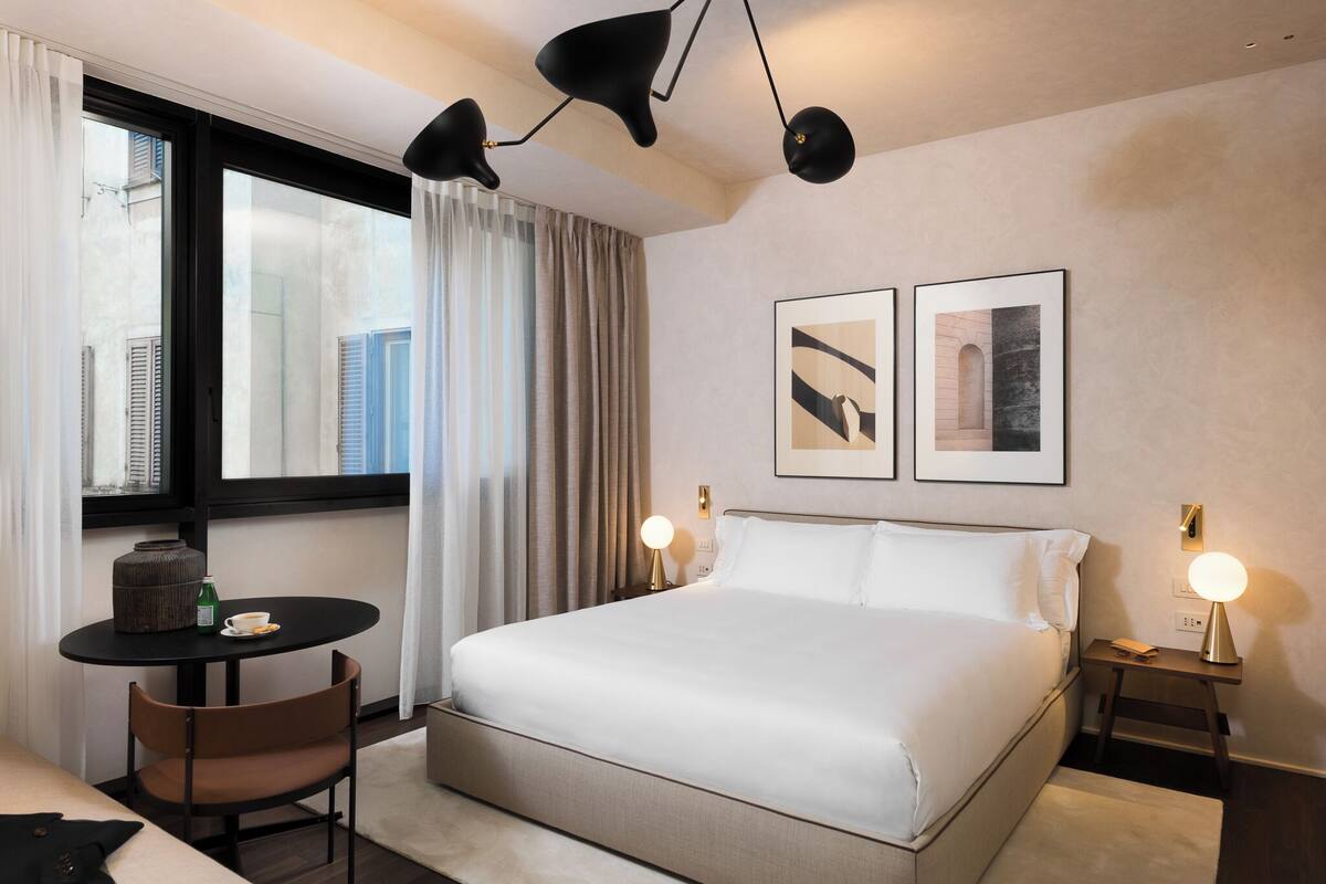 A modern hotel room with a large bed, a small round table with a chair, a desk, and a window with curtains.