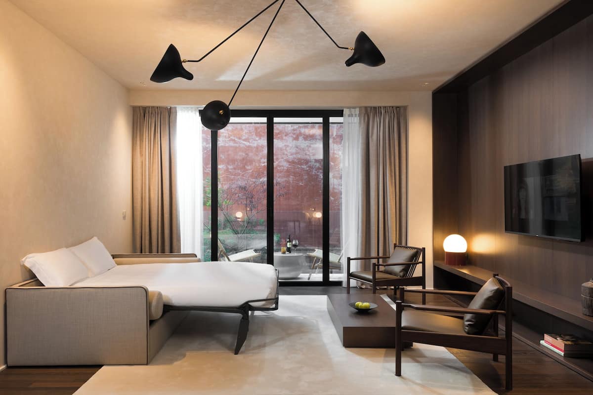A modern hotel room with a bed, a desk, a chair, a TV, and a large window with curtains.
