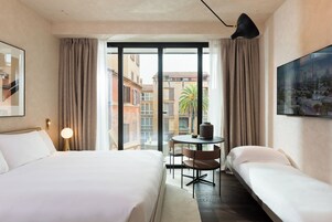 Habitación de hotel con una cama grande, otra más pequeña, una mesa redonda y vistas de edificios a través de la ventana.