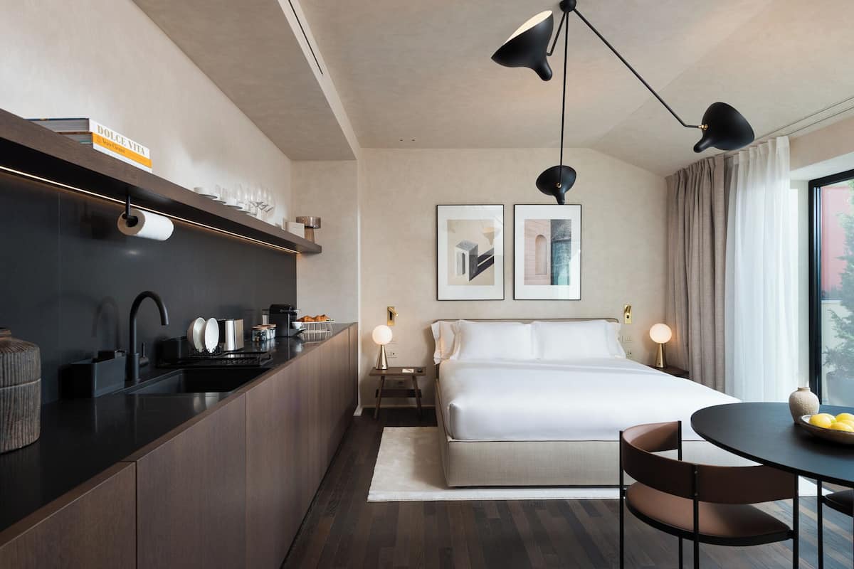 A modern hotel room with a kitchenette, a large bed, a dining area, and a view of the city.