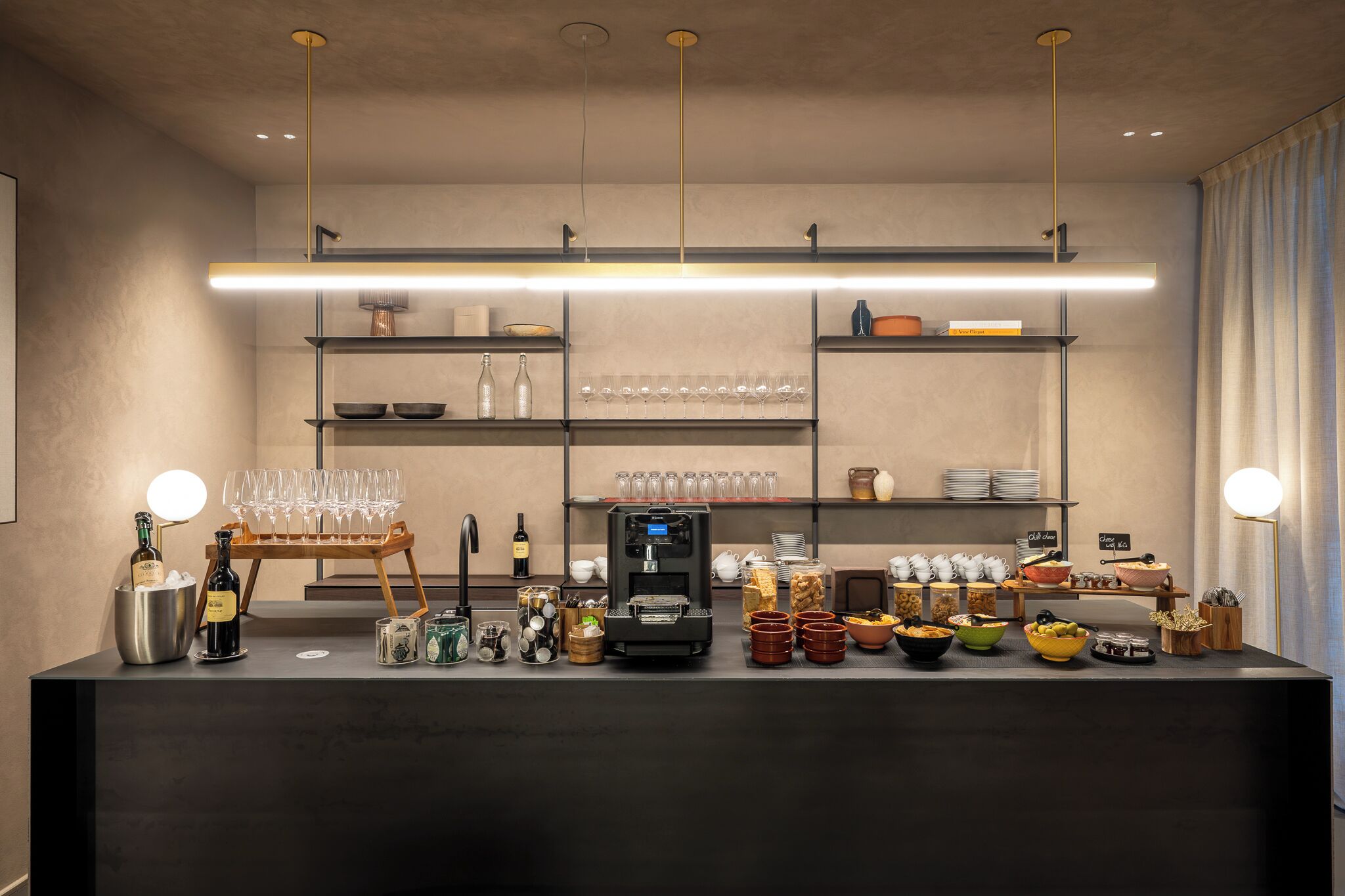 A modern bar area with a coffee machine, wine bottles, glasses, and various food items.