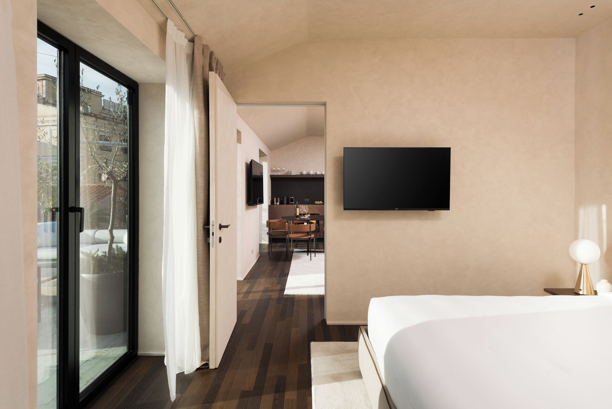 A modern hotel room with a large bed, a flat-screen TV mounted on the wall, a sliding glass door leading to a balcony, and a view of the cityscape.