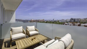 Un balcón con dos sillas blancas acolchadas y una mesa de madera, con vistas a un paisaje urbano con edificios y cielo despejado.