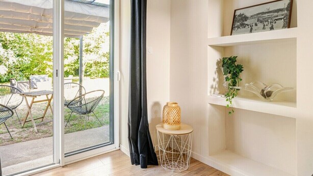 A room with a sliding glass door leading to an outdoor area with chairs and a table. There is a shelf with decorative items and a framed picture. - Charming house with garden near Lyon, airport and Eurexpo (Jonage)