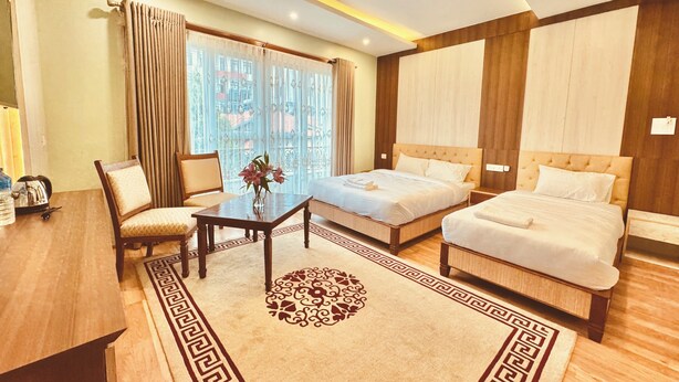 A hotel room with two beds, a wooden floor, a small table with a vase of flowers, and a view of buildings through large windows. - Hotel Bayberry Kathmandu - Near Tribhuvan International Airport (Kathmandu)