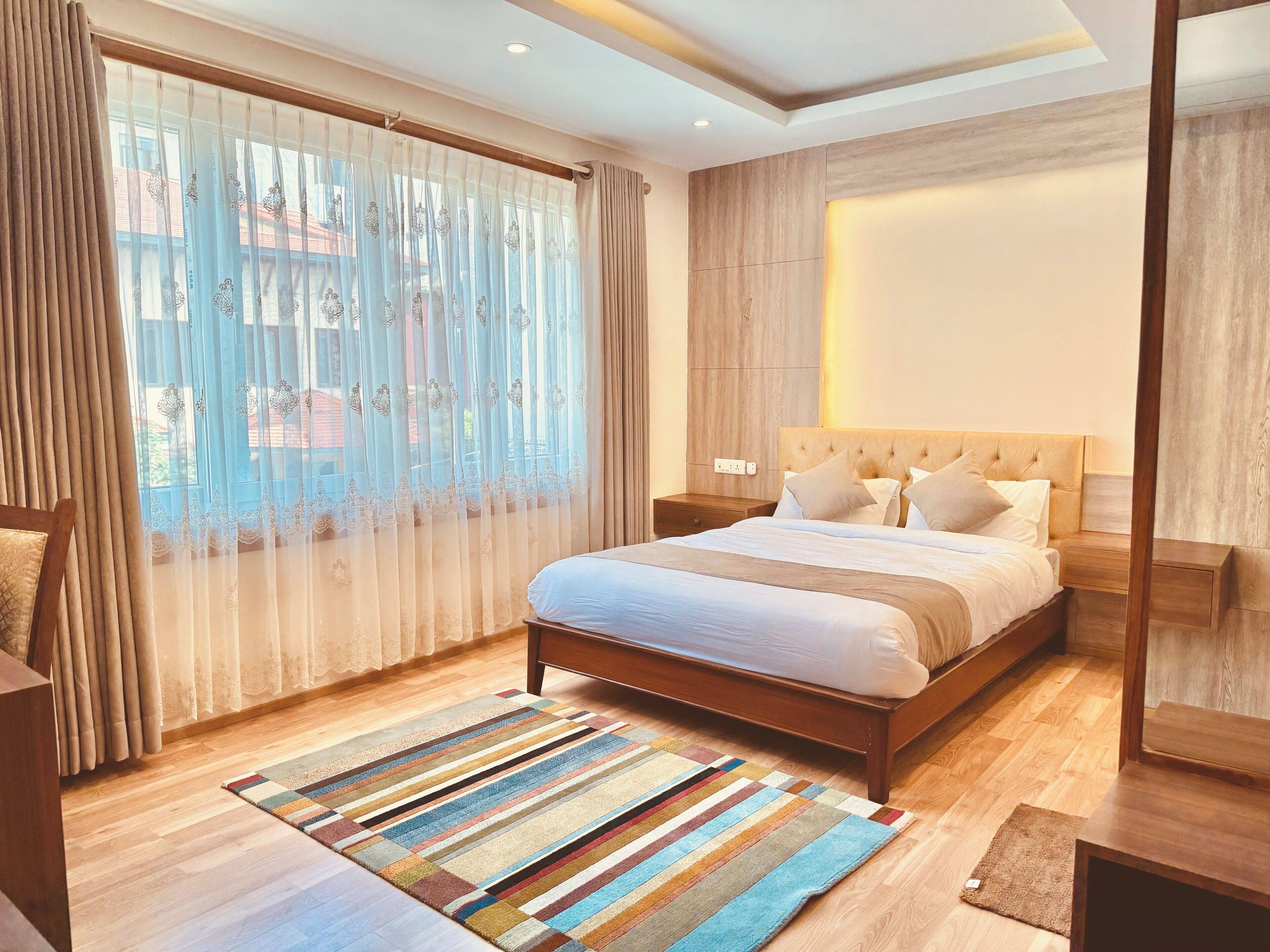 A bedroom with a bed, wooden headboard, bedside tables, a striped rug, and sheer curtains.