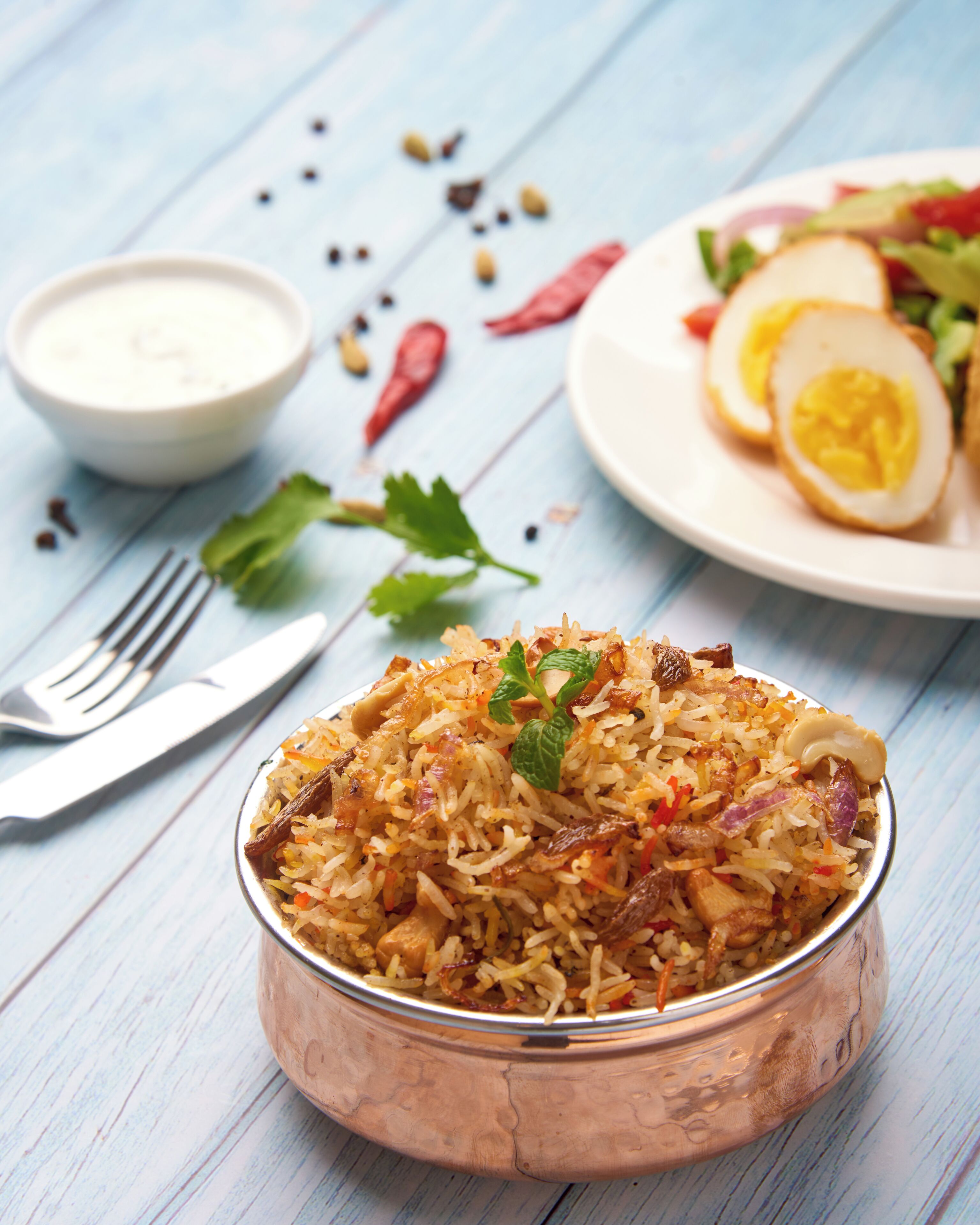 A bowl of biryani with a side of salad and a small bowl of sauce on a wooden table.