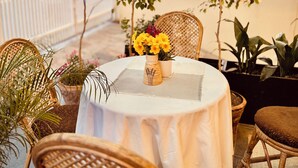 A round table with a white tablecloth, surrounded by wicker chairs, in a covered outdoor area with potted plants and a glass ceiling. - Hotel Bayberry Kathmandu - Near Tribhuvan International Airport (Kathmandu)
