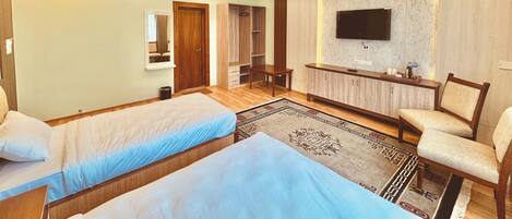 Habitación de hotel con dos camas individuales, una zona de estar con sofá y sillones, un televisor montado en la pared y un armario.
