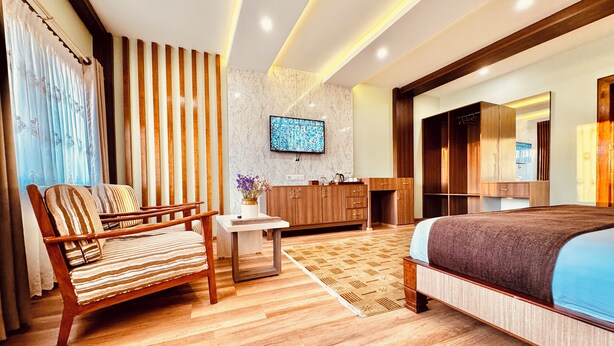 A modern hotel room with a wooden sofa, a small table with a vase, a large bed, a wooden wardrobe, a television on the wall, and a patterned rug. - Hotel Bayberry Kathmandu - Near Tribhuvan International Airport (Kathmandu)