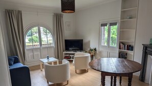 A living room with a round wooden table, two white armchairs, a blue sofa, a TV, and a bookshelf. - Apt with garden and parking on the banks of the Seine in front of the cathedral (Rouen)