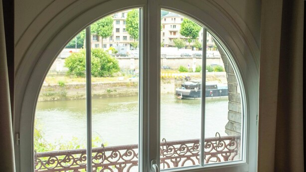 A view through a window with a decorative iron railing, overlooking a river with boats and buildings in the background. - Apt with garden and parking on the banks of the Seine in front of the cathedral (Rouen)