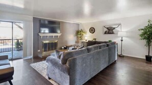 A modern living room with a gray sofa, a fireplace, a dining area, and a large window. - Hoff Gardens 39 - 2BR 2BA - Downtown Lubbock (Lubbock)