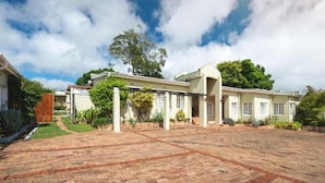 A spacious courtyard with a brick pathway leading to a building with multiple windows and a covered walkway. - Oak Lodge Guesthouse (Makhanda)
