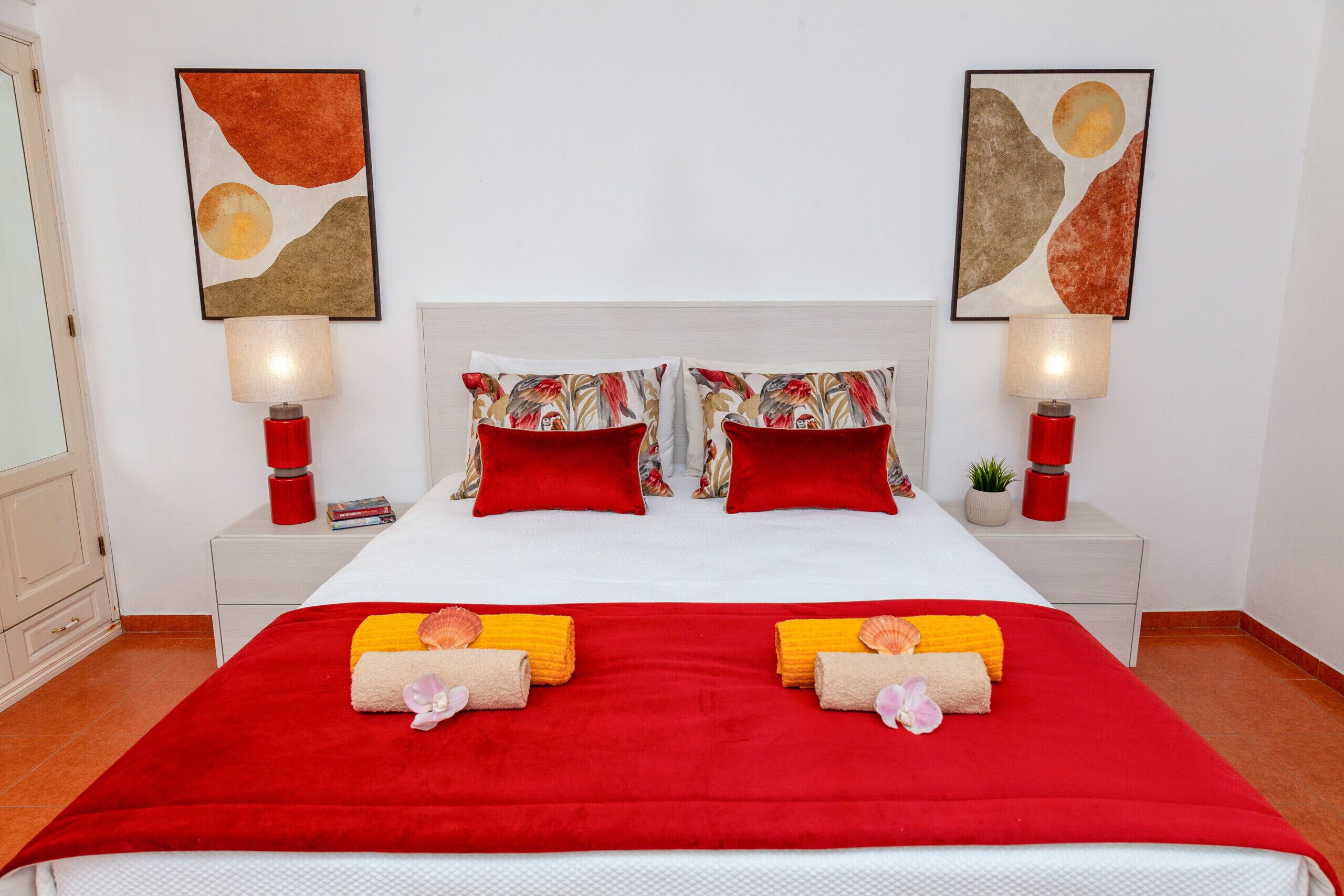 Quarto de hotel com uma cama grande, roupa de cama vermelha e branca, duas luminárias vermelhas e duas pinturas abstratas nas paredes.