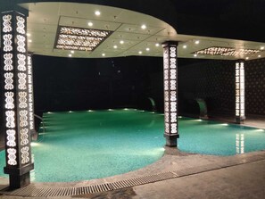 Une piscine dotée d’une zone abritée, de colonnes ornées de luminaires décoratifs et entourée d’un espace sombre.