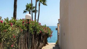 A narrow pathway flanked by a fence with flowering plants and palm trees, leading to a beach with the sea in the background. - Coastal Retreat-sleeps 8, Sea View, Pool & Parking (Estepona)