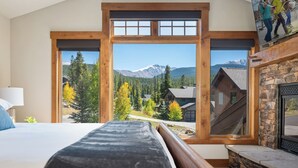 Un dormitorio con un ventanal grande, una cama, una chimenea de piedra y vistas a una montaña y un bosque.