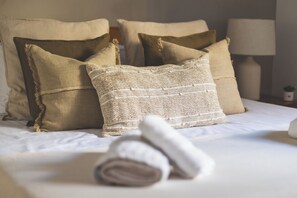 A neatly made bed with beige and white bedding, several pillows, and a rolled towel.