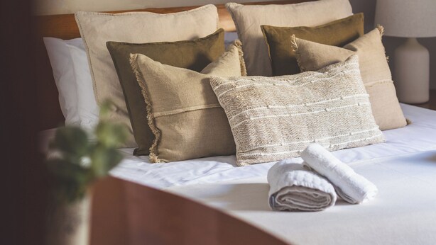 A neatly made bed with beige and textured pillows, a rolled towel, and a framed picture on the wall.