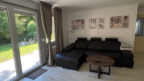A spacious living room with a sectional sofa, a round coffee table, and a large sliding glass door leading to a garden. - Newly adapted vacation apartment in the heart of Gemünd (Schleiden)