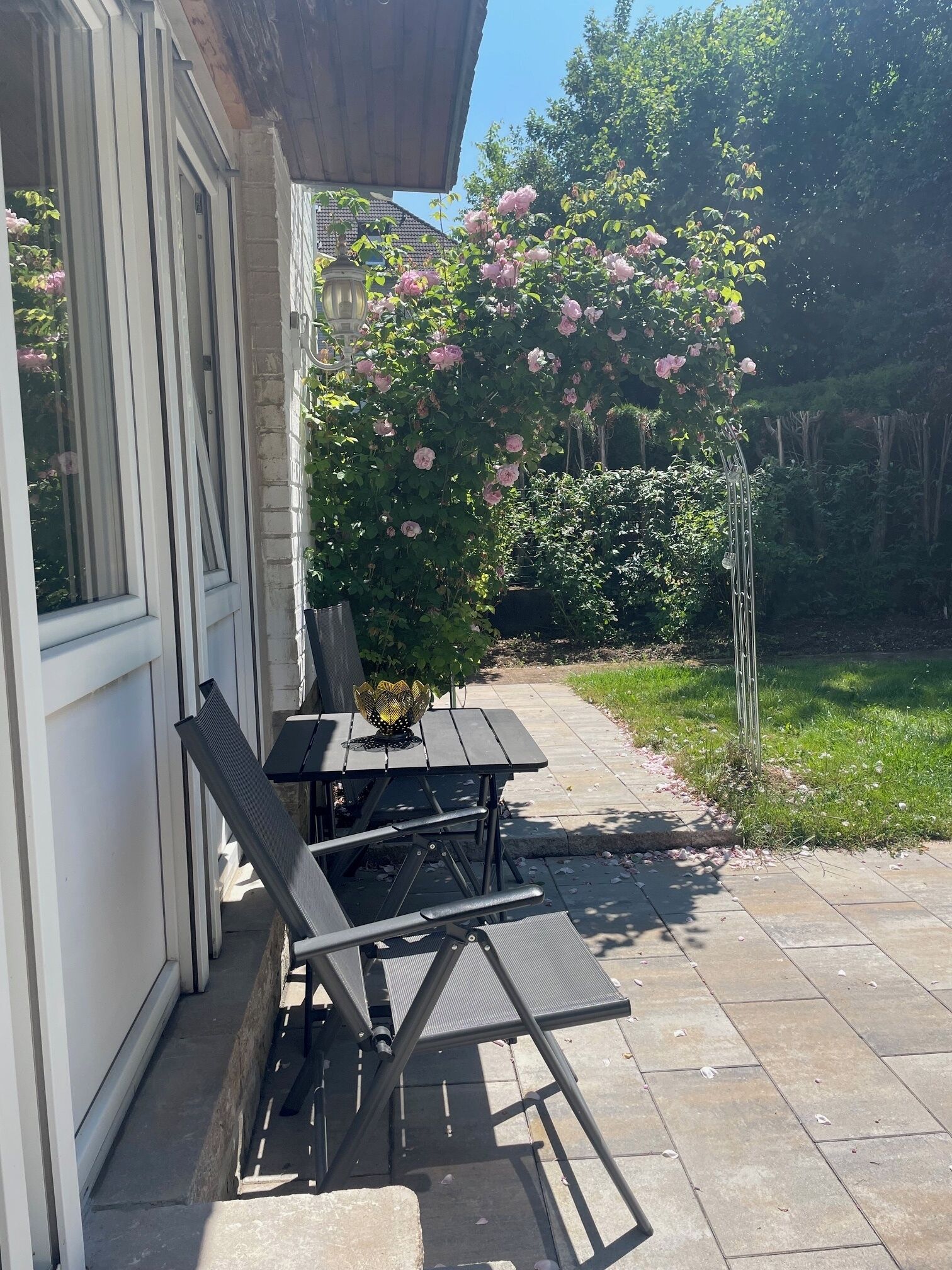 Een terras buiten met een tafel en stoelen, een tuin met roze bloemen en een pad naar een huis.