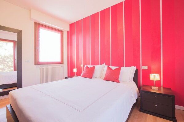 A bedroom with a large bed, a nightstand with a lamp, a window, and a red accent wall. - Marina Loft (Moniga del Garda)