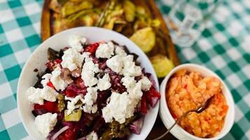 A table with a checkered tablecloth, a bowl of salad with feta cheese and red onions, a small bowl of hummus, and a tray of bread and pickles.