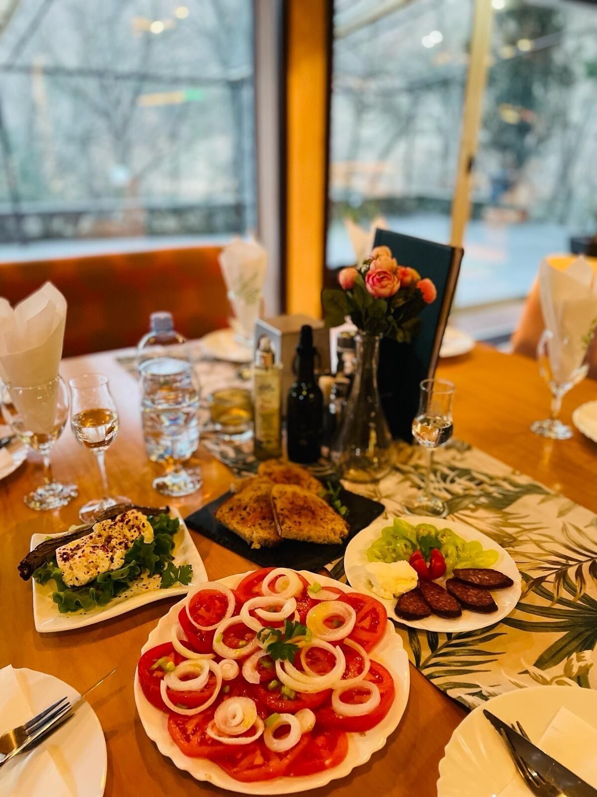 A dining table with various dishes, including a pizza with tomato and onion toppings, a plate of sliced tomatoes and onions, and a small salad. There are also wine glasses, bottles, and a vase with flowers on the table.