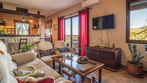 Un salon confortable avec un canapé, des fauteuils, une table basse et un téléviseur. Il y a un balcon avec des rideaux rouges et une vue sur le désert.