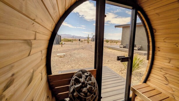 A wooden cabin with a circular window, a door, a wooden bench, and a basket of rocks. - Container Home @ Stardust Big Bend 5 min from Park (Terlingua)