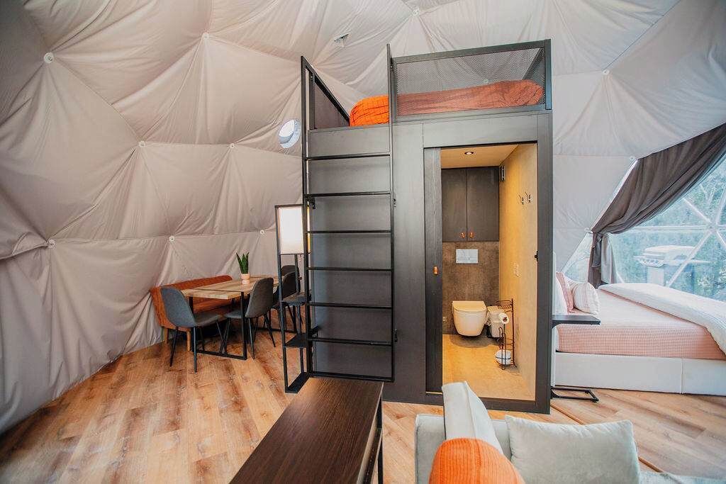 A modern tent-style accommodation with a loft bed, a dining area, and a bathroom.