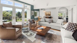 A spacious living room with a large sofa, two armchairs, and a coffee table. There is a balcony with a view of greenery and hills. - Home in Stowe with beautiful view of mountains

 (Stowe)