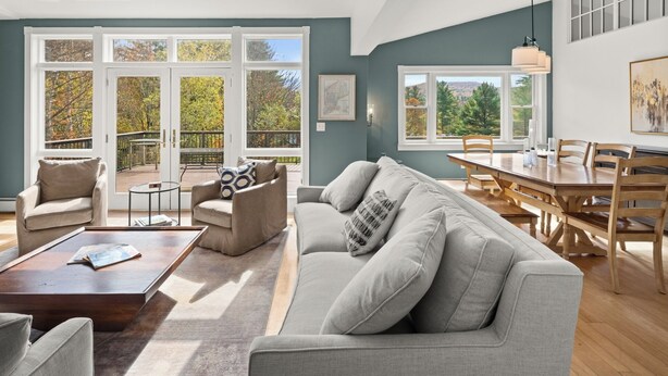 A spacious living room with a grey sofa, a coffee table, and a dining area with a wooden table and chairs. Large windows provide views of a garden and trees. - Home in Stowe with beautiful view of mountains

 (Stowe)