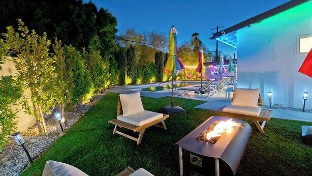 A well-lit outdoor patio area with lounge chairs, a fire pit, and umbrellas. - Navigate Stays Iconic Home w/ Salt Pool Spa Casita (Palm Springs)