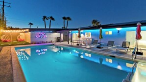 A pool area with lounge chairs and umbrellas, a neon sign reading "PALM SPRINGS", and palm trees in the background. - Navigate Stays Iconic Home w/ Salt Pool Spa Casita (Palm Springs)