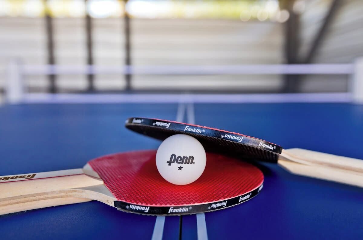 Une table de ping-pong recouverte d’un feutre rouge, une balle blanche et deux raquettes de ping-pong.