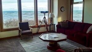 Ein Zimmer mit großen Fenstern und Meerblick, ein rotes Sofa, ein Sessel, ein Teleskop und eine Lampe.