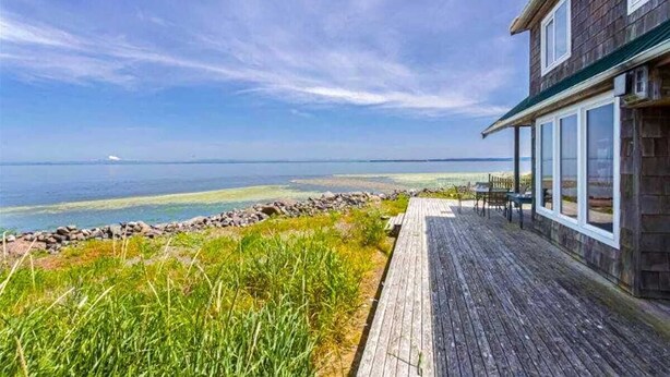 Eine Holzterrasse führt zu einem Haus mit großen Fenstern mit Blick auf Strand und Meer.