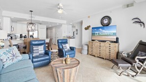 Ein modernes Wohnzimmer mit einer Couch, zwei blauen Ledersesseln, einem Couchtisch und einem Fernseher, der 'hosteeva' anzeigt.