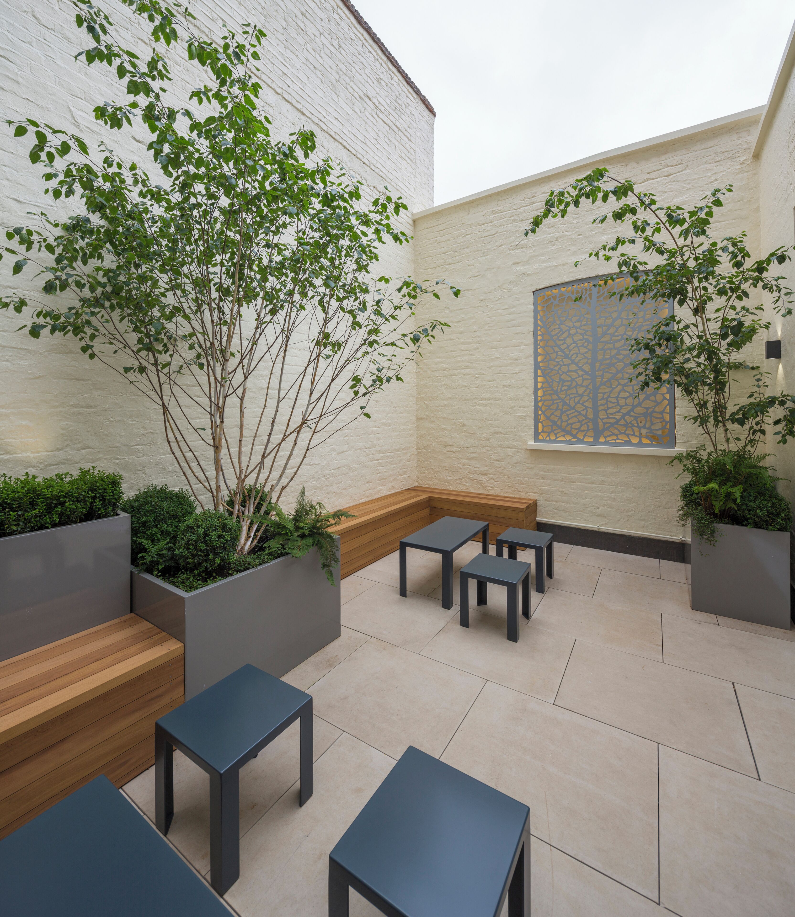 A modern outdoor seating area with wooden benches and tables, surrounded by planters and a white brick wall.